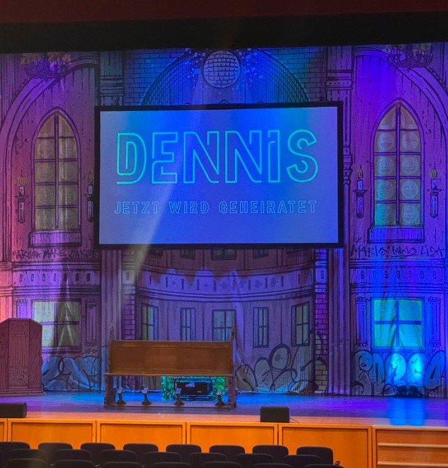 Theaterbühne mit Leinwand „Dennis – Jetzt wird geheiratet“ in blauem und violettem Licht
