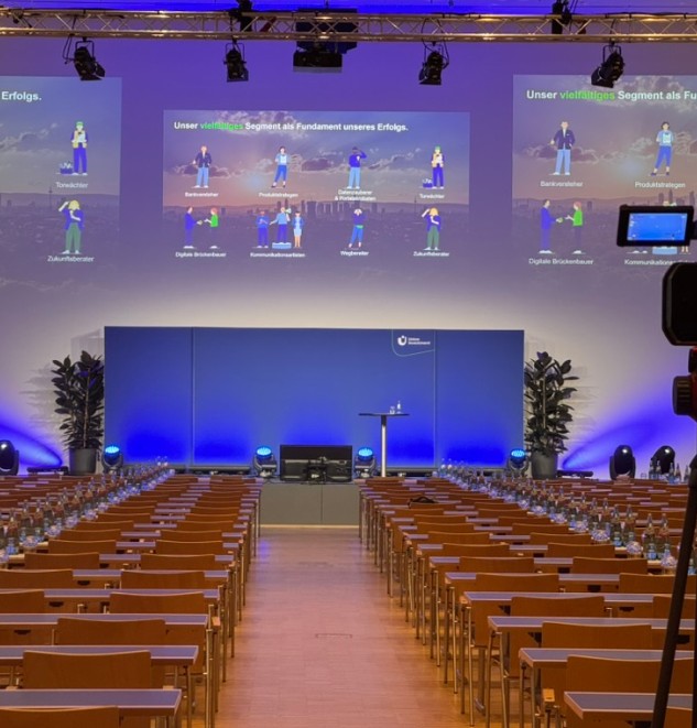 Konferenzsaal mit Stuhlreihen, blau beleuchteter Bühne und großen Projektionsflächen