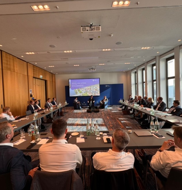 Konferenz in einem hellen Tagungsraum mit Teilnehmenden an langen Tischen und Präsentation auf dem Bildschirm